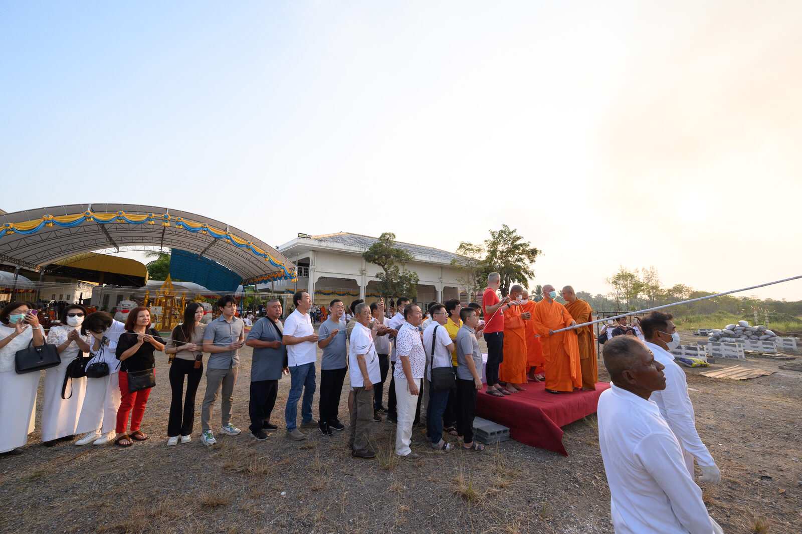 พิธีเททองกลางลานโรงงานวุ้นเส้นคลองขลุง พระสงฆ์อยู่บนเวที พุทธศาสนิกชนยืนสายสิญจน์